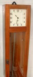 External view of Gent clock with wood case, white square deco style face and pendulum behind a glass window