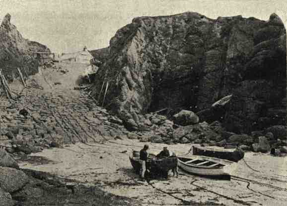 A wide track leads down between rocks to a sandy beach with fishermen working on their boats